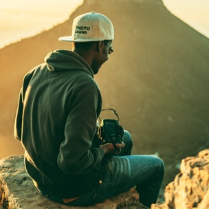 A black man sits on a large rock with his camera with large mountains in the background
